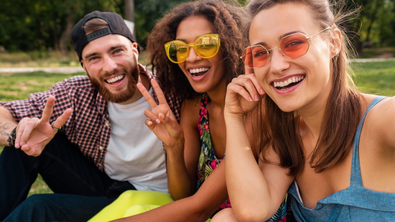 colorful stylish happy young company of friends sitting park, man and women having fun together, summer hipster fashion style, traveling with camera, taking selfie pictures, smiling and positive